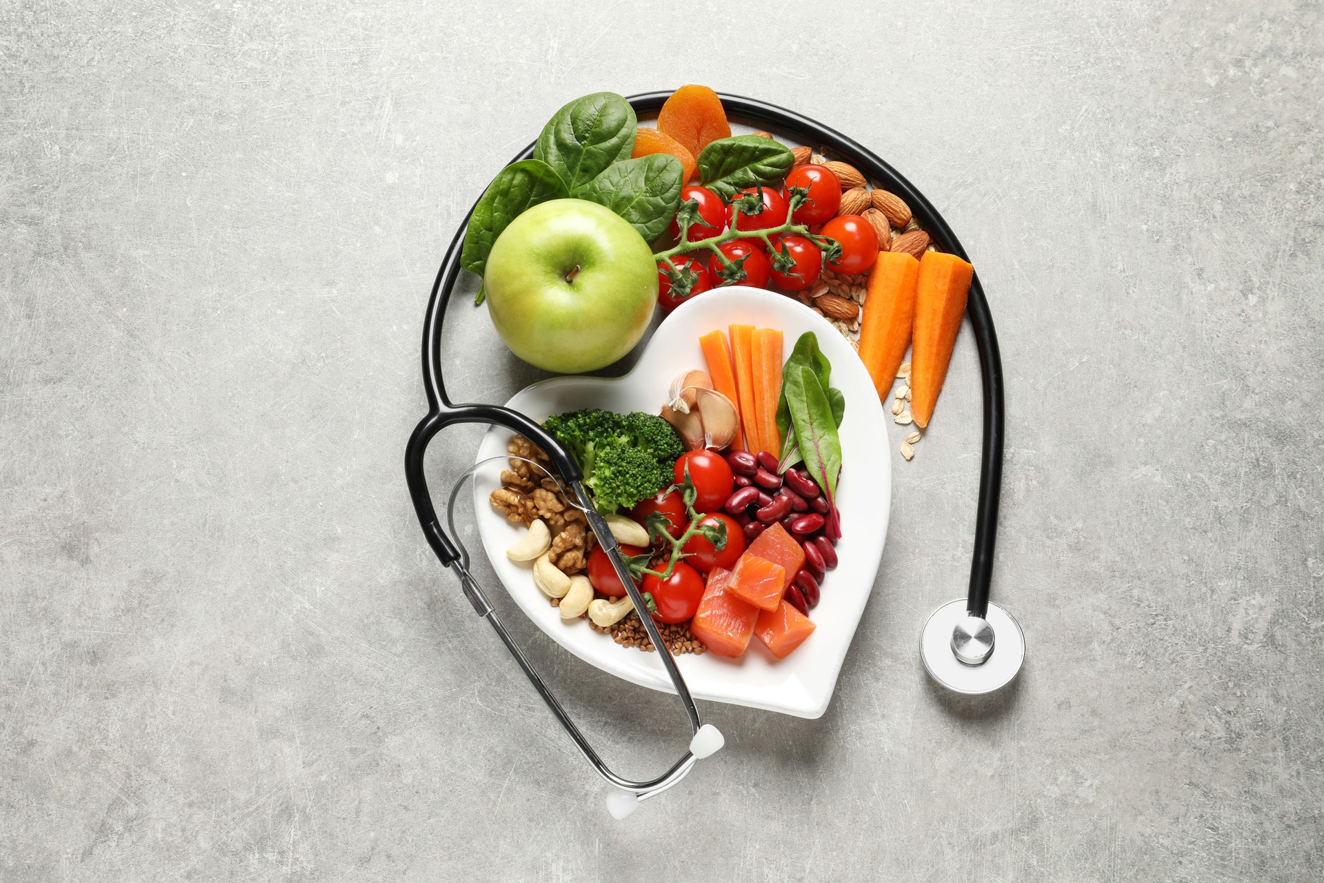 Healthy food concept photograph showing a heart shaped dish filled with vegetables, fruit, nuts, and salmon wrapped with a medical stethoscope.